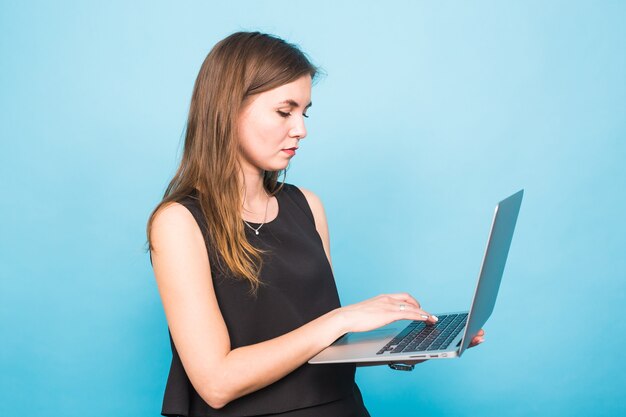 Smiling young woman standing and holding laptop over blue background.