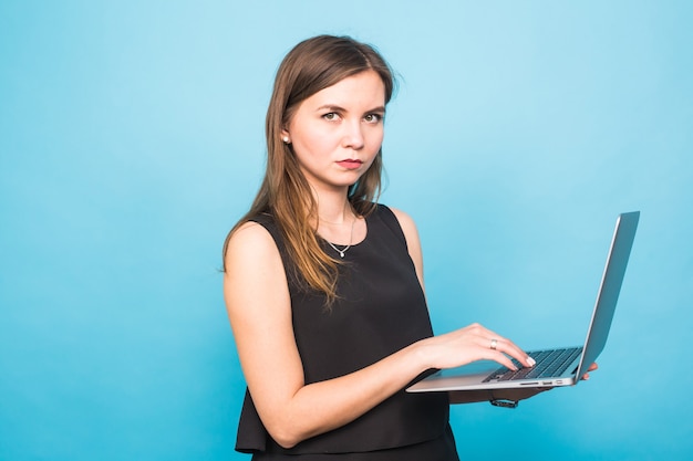 Smiling young woman standing and holding laptop over blue background.