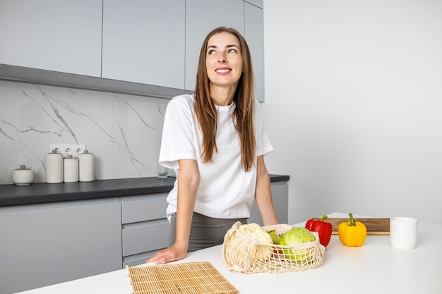 Giovane donna sorridente in piedi vicino al tavolo con verdure acquistate in borsa in cucina