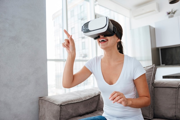 Улыбающаяся молодая женщина, стоящая и использующая очки виртуальной реальности дома