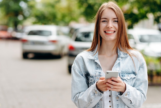 スマートフォンで立っていると屋外のカメラを見て笑顔の若い女性