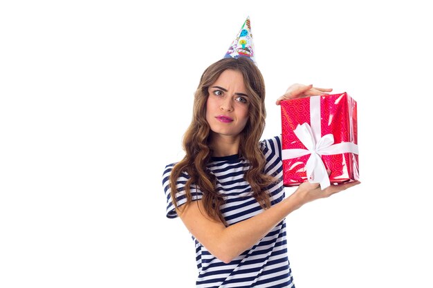 Smiling young woman in stripped Tshirt and celebration cap holding red present in studio