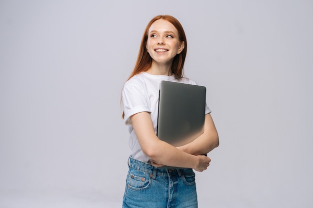 Smiling young woman student holding laptop computer and looking away on isolated gray background