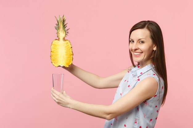 Smiling young woman in summer clothes holding half of fresh ripe pineapple fruit, glass cup isolated on pink pastel wall background. People vivid lifestyle, relax vacation concept. Mock up copy space.