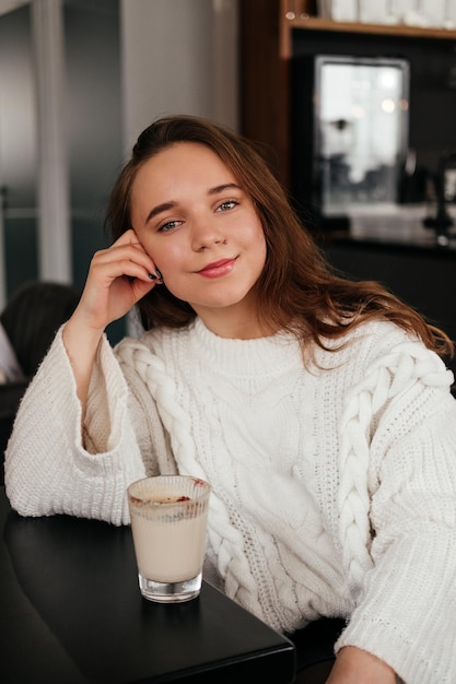 屋内カメラを見てカフェに座ってコーヒーを飲みながらセーターで笑顔の若い女性