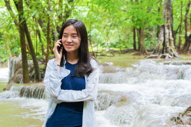 Smiling young woman talking over phone while standing against waterfall and trees