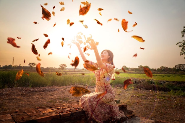 Smiling young woman throwing dry leaves while sitting against sky during sunset
