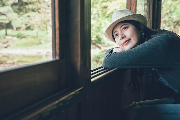 smiling young woman traveler with head lean through the open window of wooden house looking up to sky daydreaming. beautiful lady putting head on hands enjoy view of green garden in japanese style.