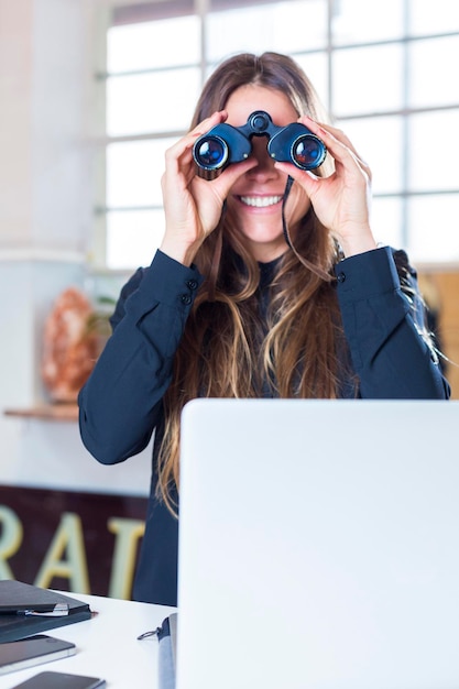 Smiling young woman using binoculars and laptop at desk