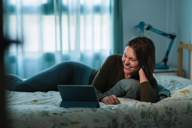 Photo smiling young woman using digital tablet while lying on bed at home