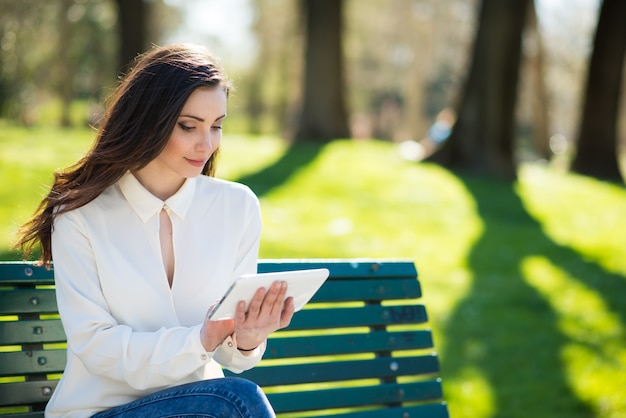 Улыбается молодая женщина, используя ее планшет в парке