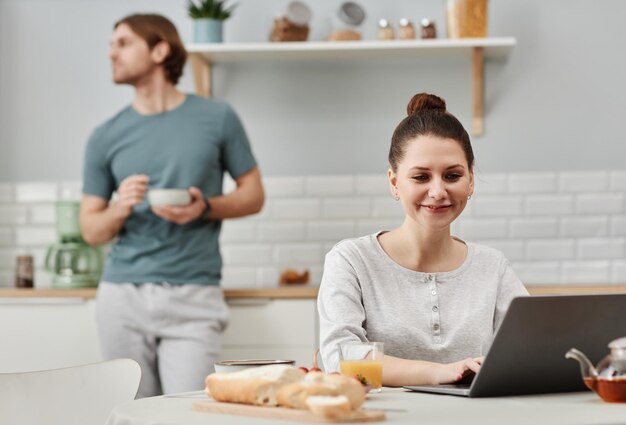 Smiling young woman using laptop in kitchen during breakfast