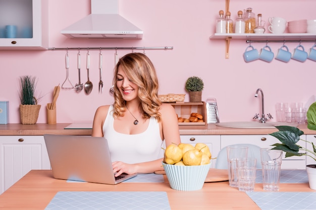 Giovane donna sorridente che utilizza computer portatile nella cucina a casa. la donna bionda lavora al computer, alle free lance o al blogger che lavora a casa