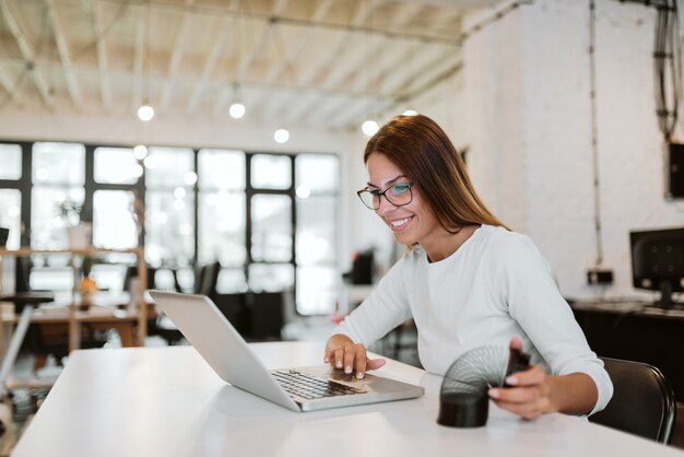 Smiling young woman using laptop and playing with slinky in bright office.