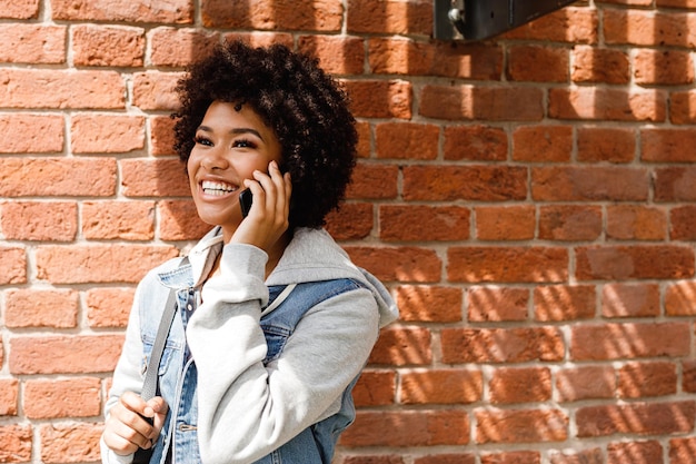 Photo smiling young woman using phone against brick wall