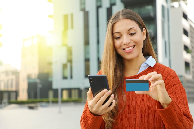 Photo smiling young woman using phone and credit card while standing in city