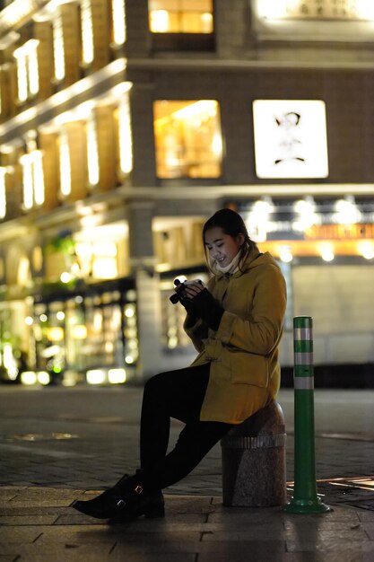 写真 夜ボラードに座っている間電話を使っている笑顔の若い女性