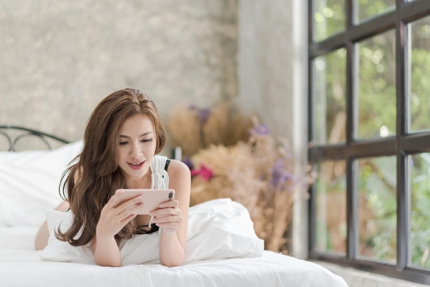 家でベッドに横たわってスマートフォンを使っている笑顔の若い女性