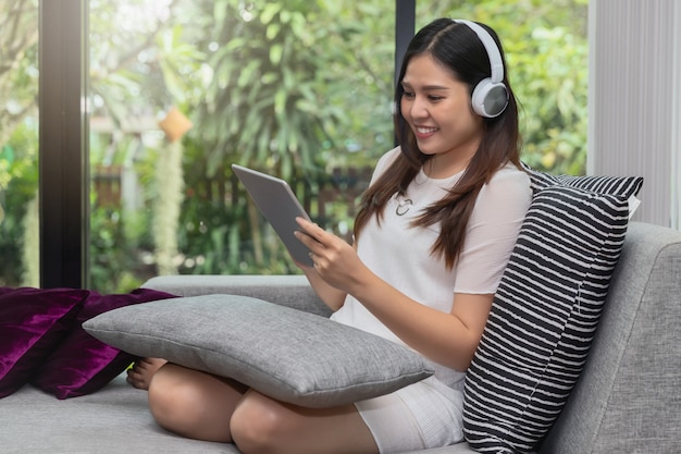 笑顔の若い女性は、ソファに座っているとタブレットを使用してヘッドフォンを着用します。