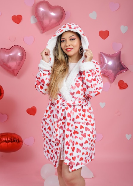 Smiling young woman wearing bathrobe with red hearts Pink background with balloons and hearts