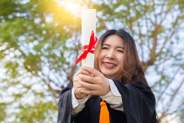 写真 卒業式のドレスを着た笑顔の若い女性が木の前で卒業証書を握っています