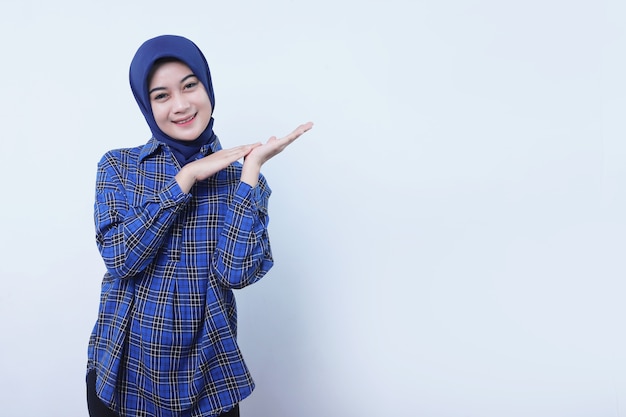 Smiling young woman wearing hijab showing something over hands isolated on white wall