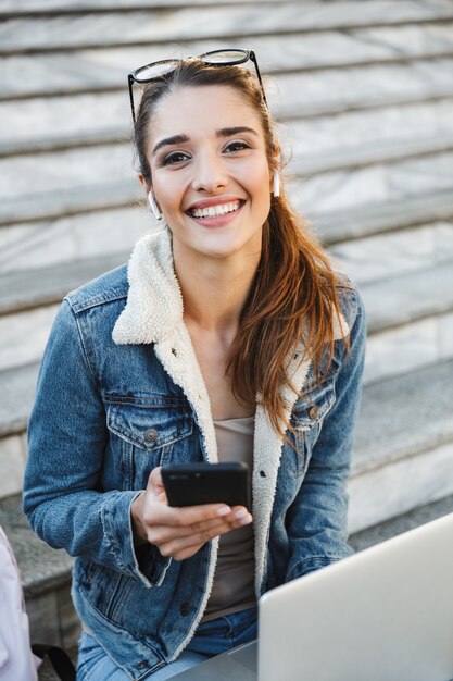 Smiling young woman wearing jacket sitting on stairs outdoors, using laptop computer, holding mobile phone