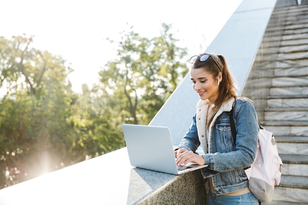 Smiling young woman wearing jacket standing at the stairs outdoors, using laptop computer