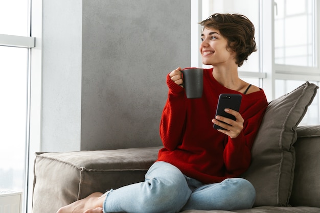 自宅のソファでリラックスしたセーターを着て、コーヒーを飲みながら笑顔の若い女性