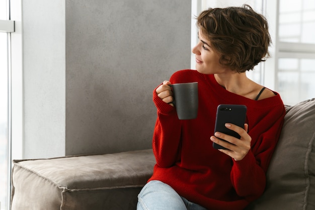 Smiling young woman wearing sweater relaxing on a couch at home, drinking coffee