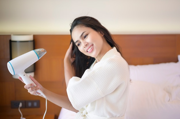 Smiling young woman wearing white bathrobe drying her hair with a hairdryer after a shower in bedroom

