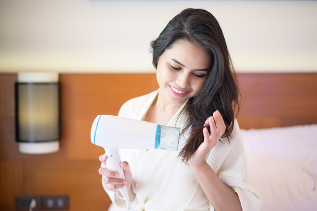 Smiling young woman wearing white bathrobe drying her hair with a hairdryer after a shower in bedroom

