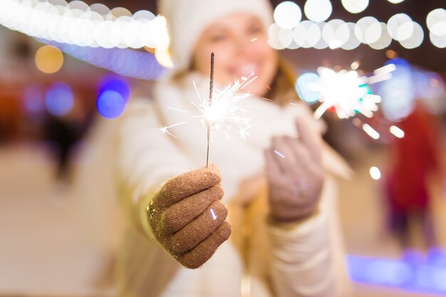 雪の背景の上に屋外で線香花火を保持している冬のニットの服を着て笑顔の若い女性。クリスマス休暇。
