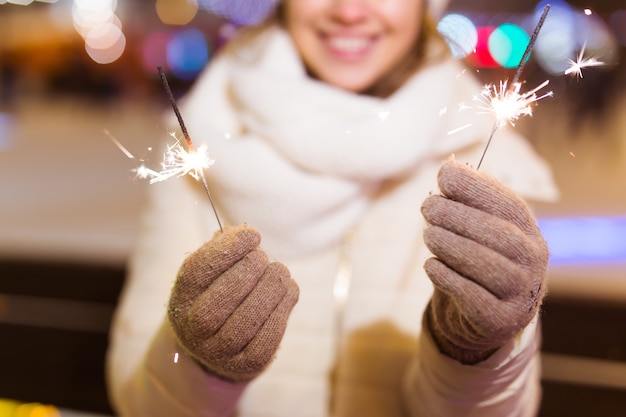 雪の背景の上に屋外で線香花火を保持している冬のニットの服を着て笑顔の若い女性。クリスマス休暇。