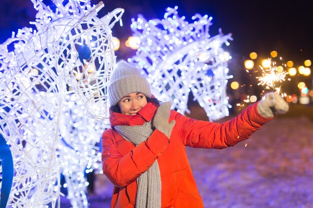 雪の背景の上に屋外で線香花火を保持している冬のニット服を着て笑顔の若い女性