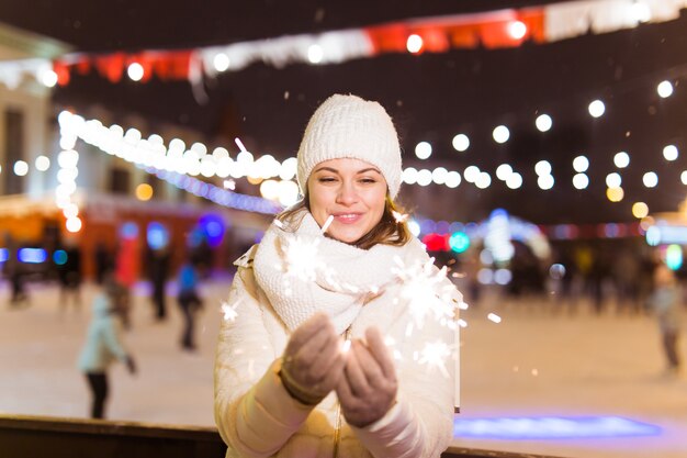 Smiling young woman wearing winter knitted clothes holding sparkler outdoors over snow background