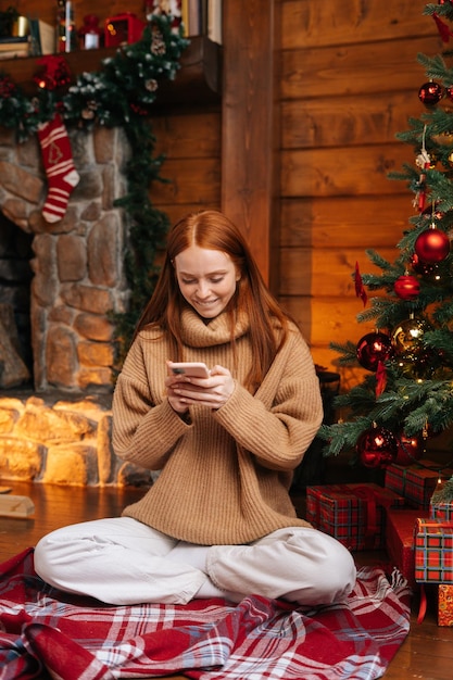 Smiling young woman wearing winter sweater using mobile phone sitting on floor near Christmas tree