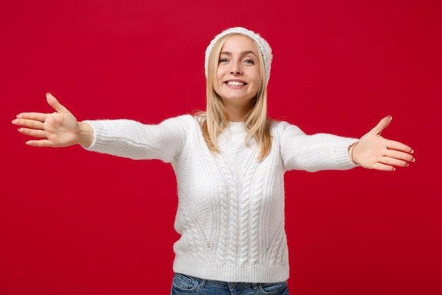 Smiling young woman in white sweater, hat isolated on red background, studio portrait. Healthy fashion lifestyle, cold season concept. Mock up copy space. Standing with spreading outstretched hands.