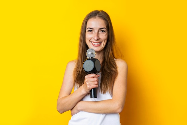 Smiling young woman in white tank top holding microphone on yellow background