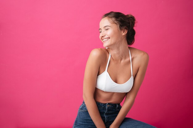 Smiling young woman in white top and jeans sitting and looking away isolated on the pink wall