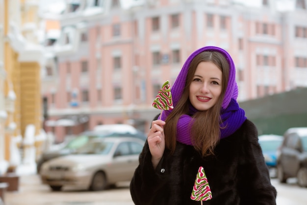 Smiling young woman in winter clothes posing with tasty colorful candies at the street