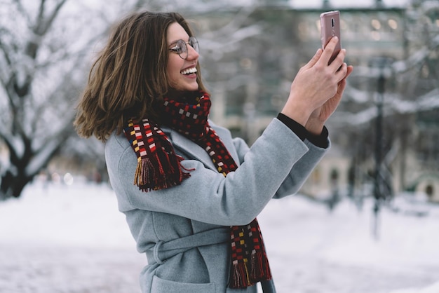 雪のある通りでスマートフォンでビデオ通話を持っている冬の衣装で笑顔の若い女性