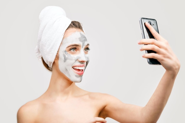 Smiling young woman with applied clay mask and towel on head sending funny selfie
