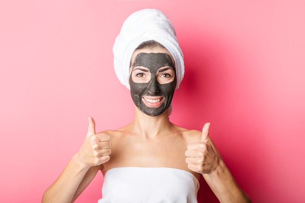 Smiling young woman with black face mask on pink background showing thumbs up gesture.