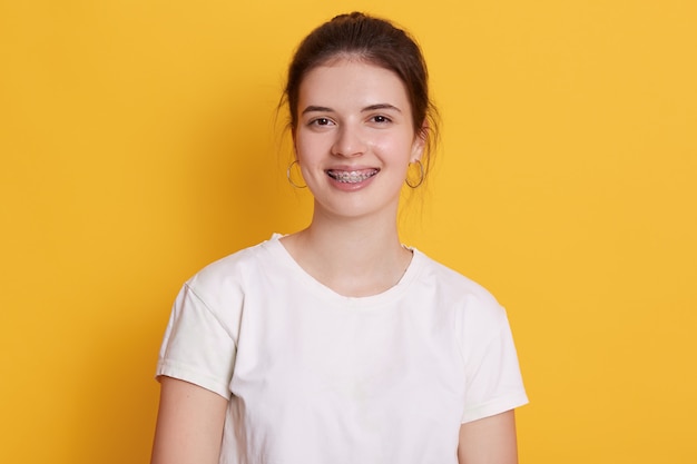 Photo smiling young woman with brackets and rounded earrings posing against yellow wall