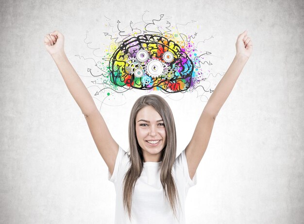 Smiling young woman with brown hair wearing a white t shirt is standing with her hands in the air and looking at the viewer. A concrete wall background with a cog brain sketch on it.