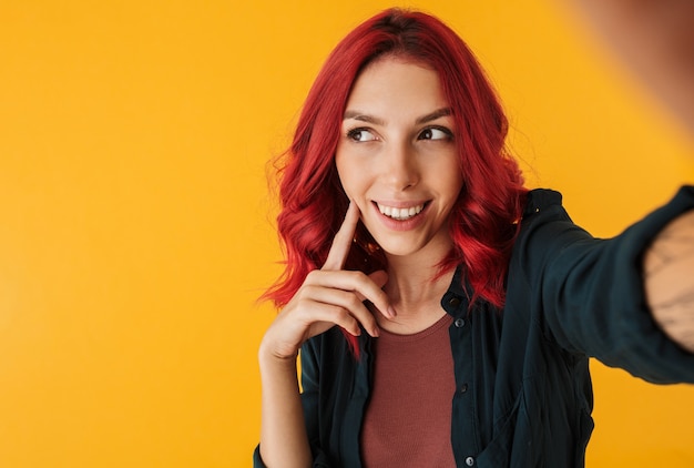 smiling young woman with curly red hair looking aside and taking selfie photo isolated