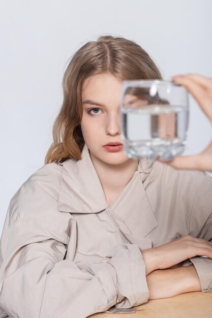 Smiling Young Woman with glass of Water, beautiful girl in beige raincoat posing in Studio