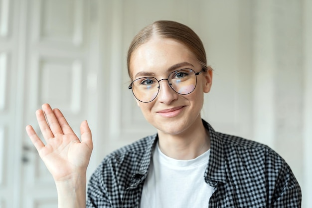 Smiling young woman with glasses greeting with a wave in a bright airy room