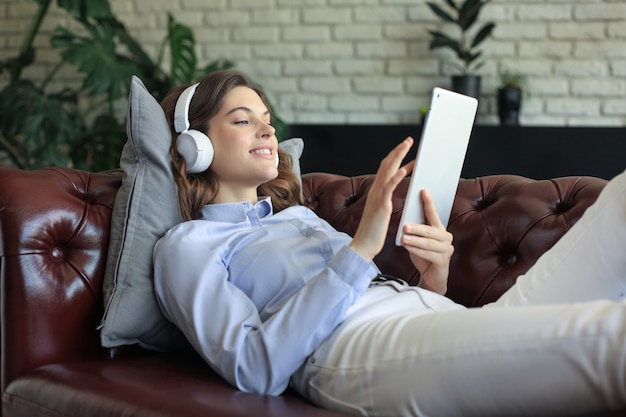 Smiling young woman with headphones and digital tablet on the sofa.